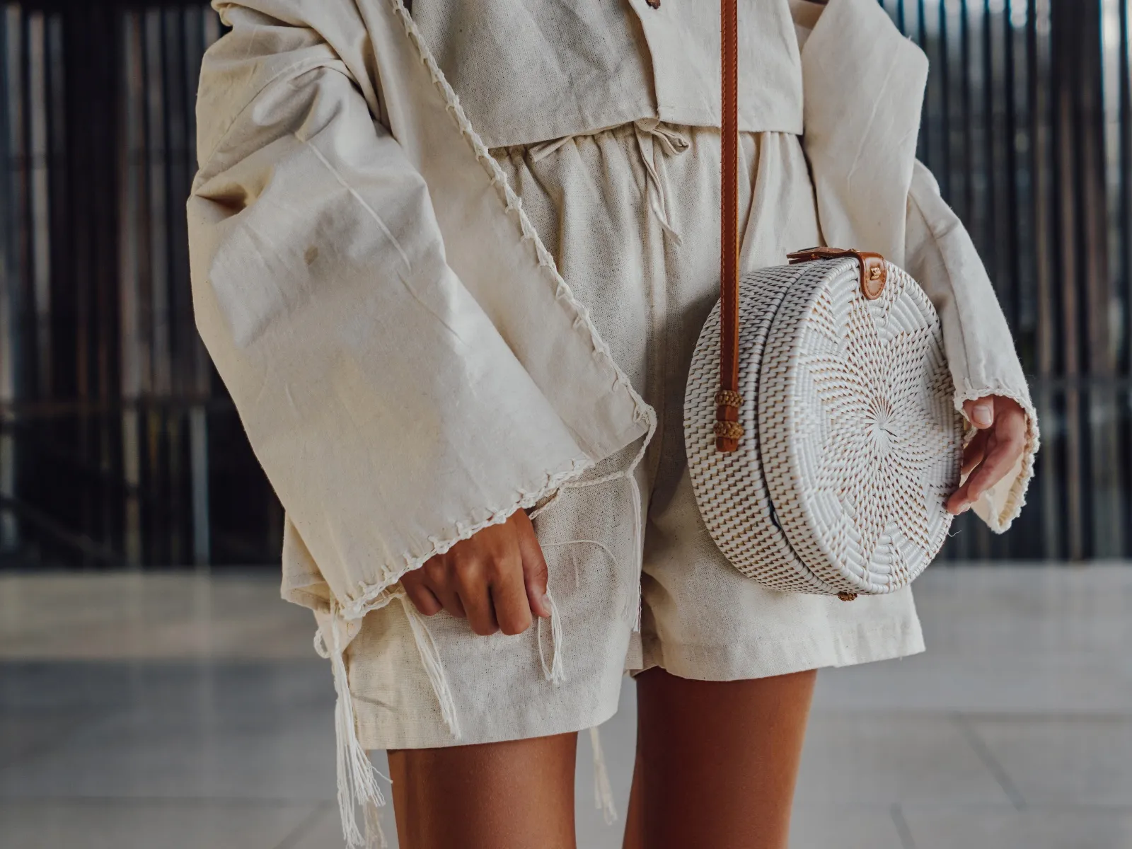 Dettaglio di un completo in lino color crema, con giacca ampia e pantaloncini, e una borsa tonda di rattan a tracolla.