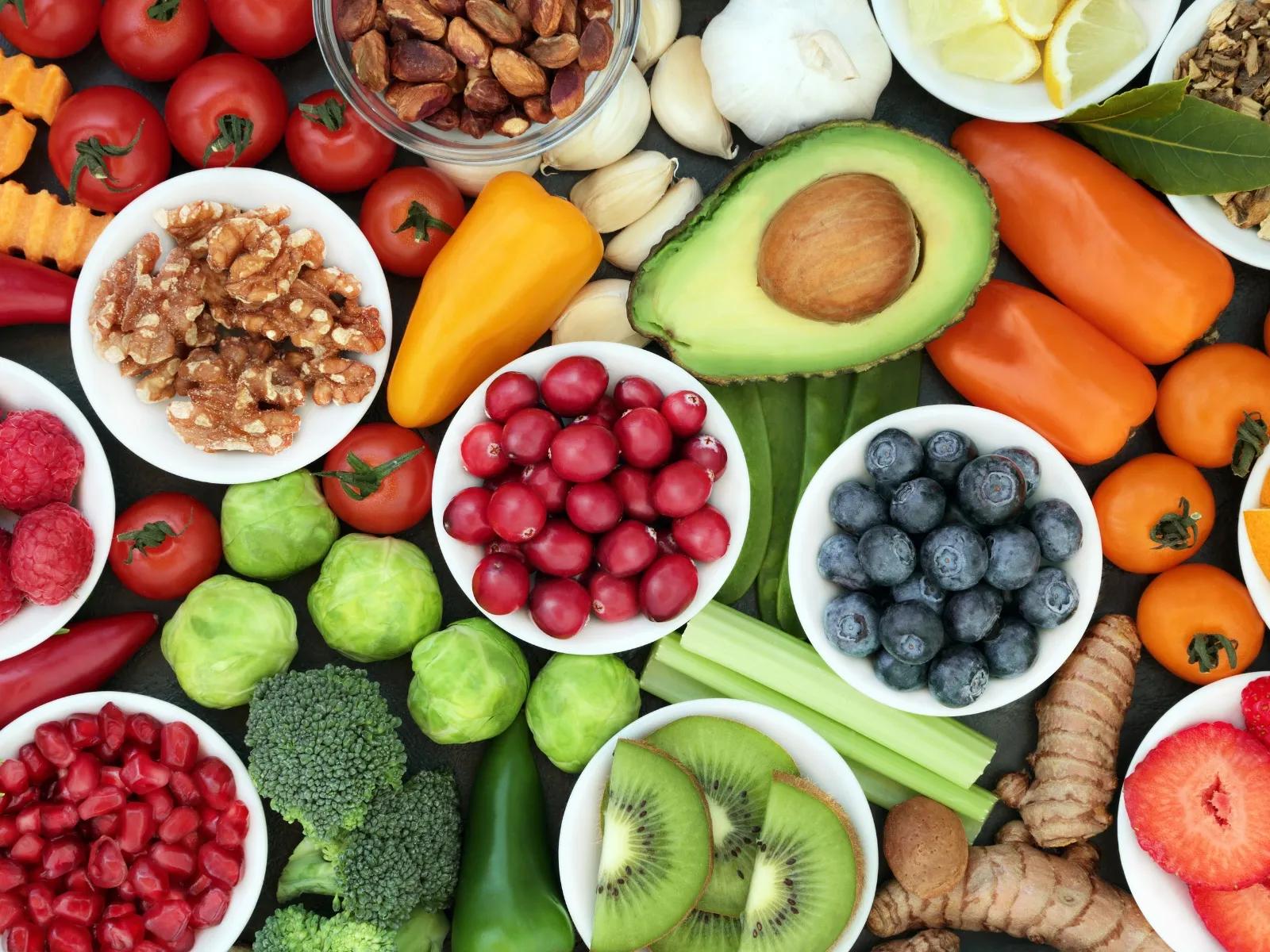 Frutta, verdura e frutta secca su un piano scuro: avocado, mirtilli, kiwi, broccoli, pomodorini e noci in ciotole bianche.