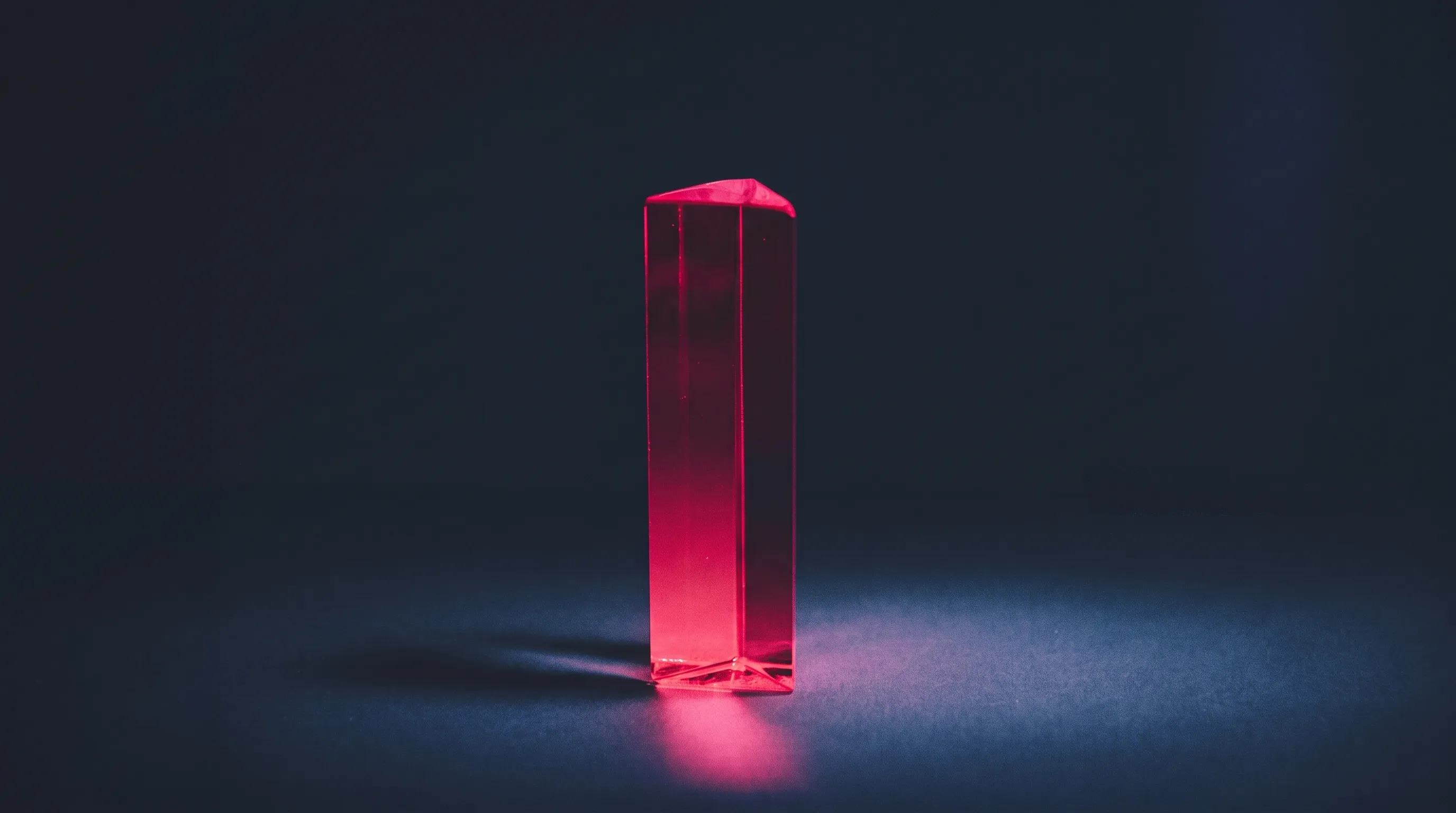 Un prisma traslucido fucsia su una superficie blu scuro, con un riflesso magenta e una lunga ombra.